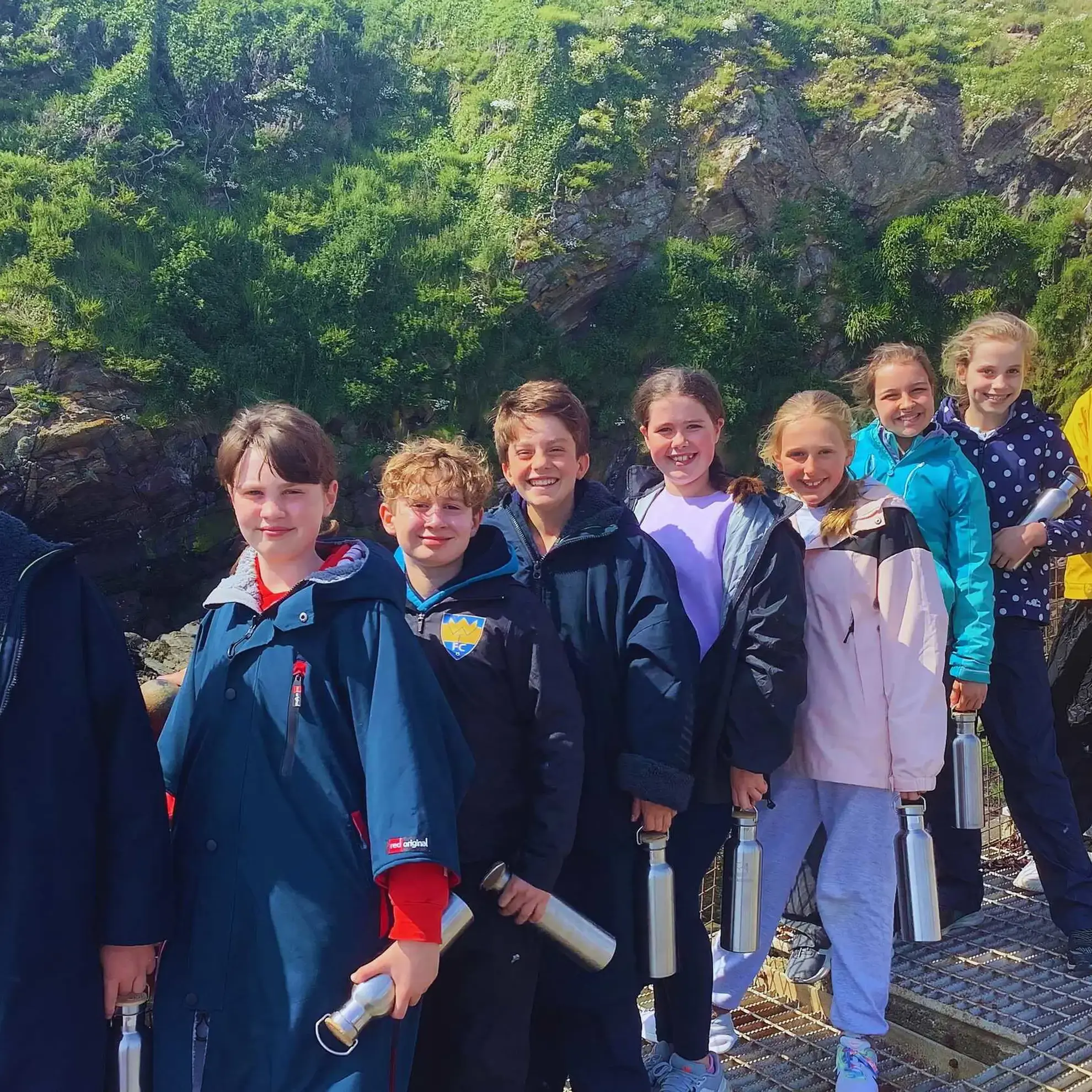 Students pose for a picture on a bridge, with grass-covered rock face in the background