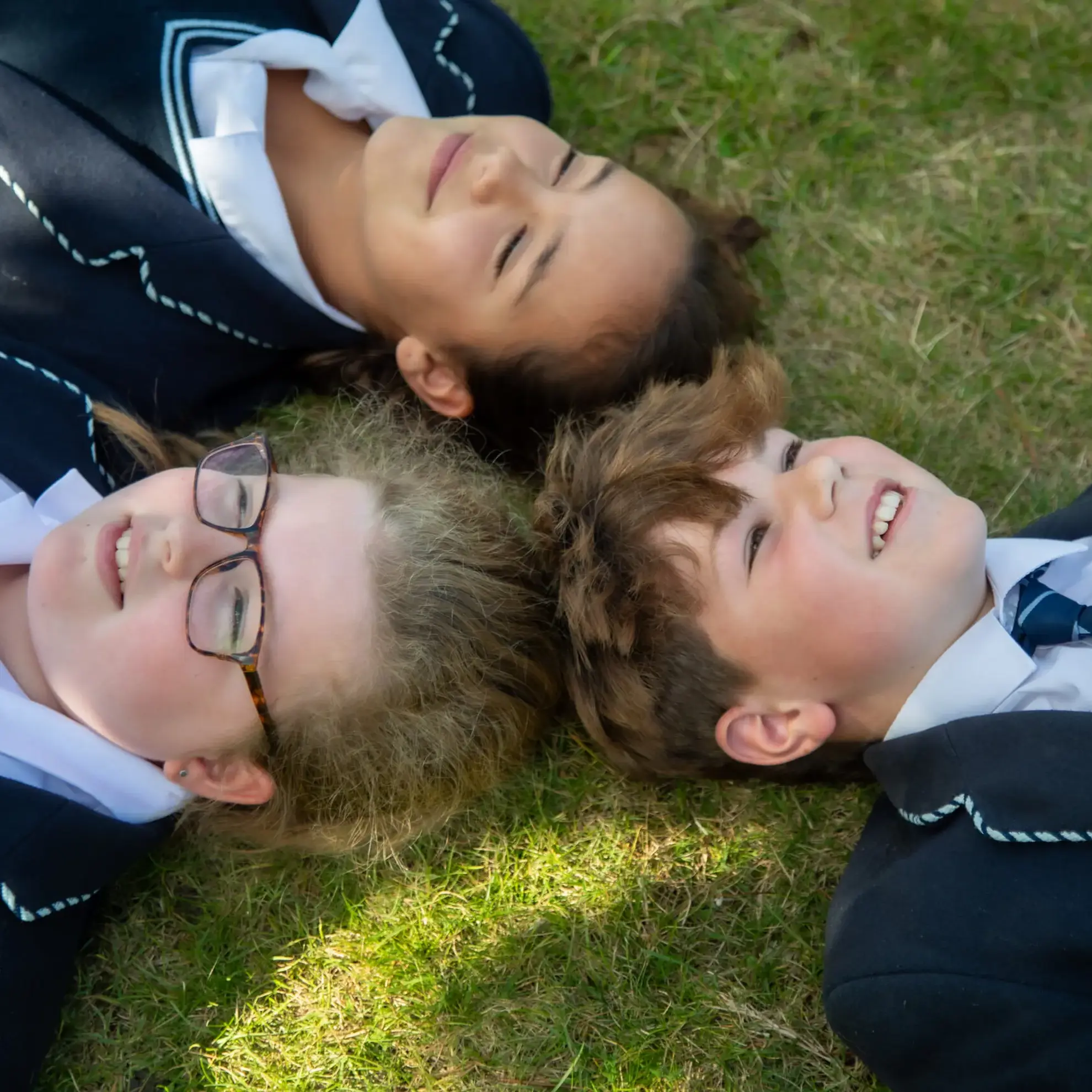 Three students lying head-to-head on grass, looking up at the sky